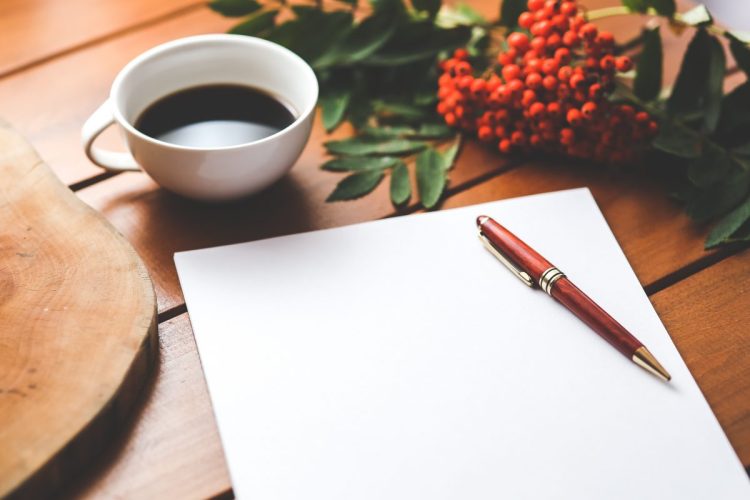 A wooden table top with blank paper and a pen and black coffee in a white mug.