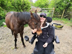 A girl (Holly) in a powered wheelchair in a field, with a bay pony stood next to her with her head nuzzling into the girl. The girl is wearing a black cap and dark glasses, a black coat and leggings.
