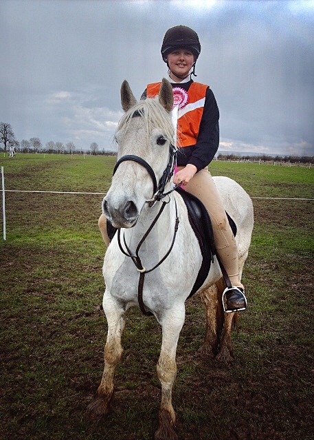 Mounted Games. A girl on a grey fell pony wearing an orange bib and a pink rosette, stood in a muddy field.