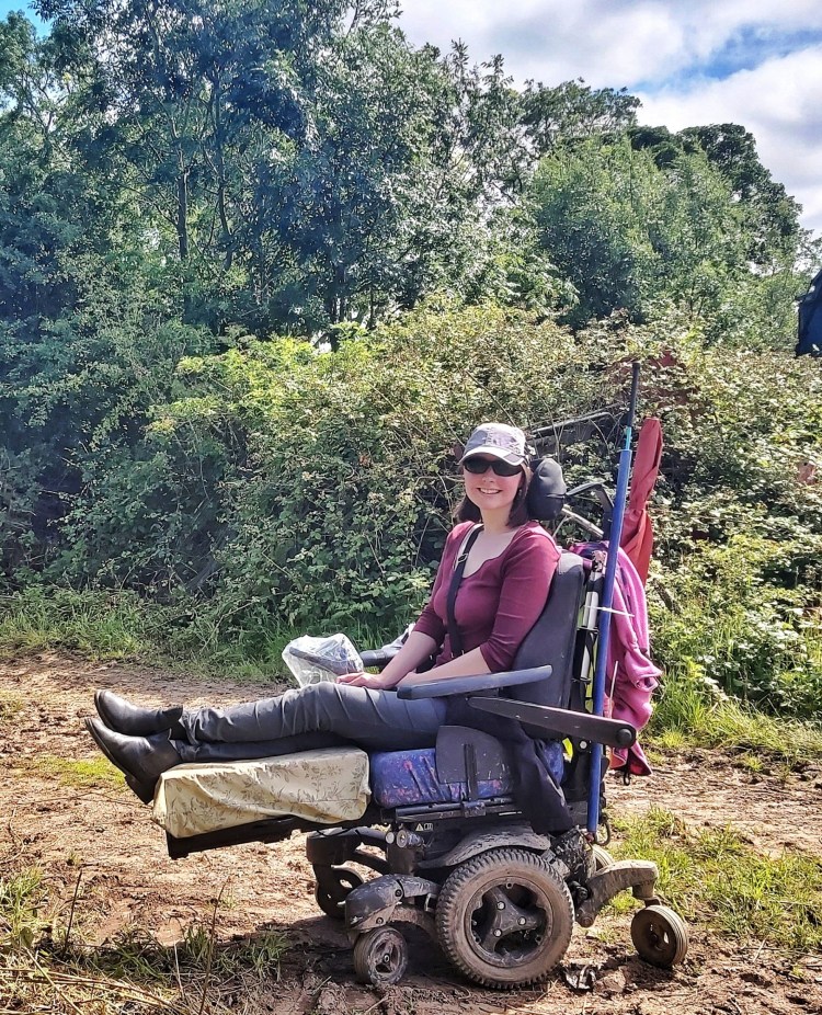 Holly, a white woman with shoulder length brown hair, sitting outside in front of hedges and trees in a Quickie Q700 Sedeo Pro Powered Wheelchair with the legs elevated. Wearing grey trousers, a plum coloured top, sunglasses and a grey baseball cap.