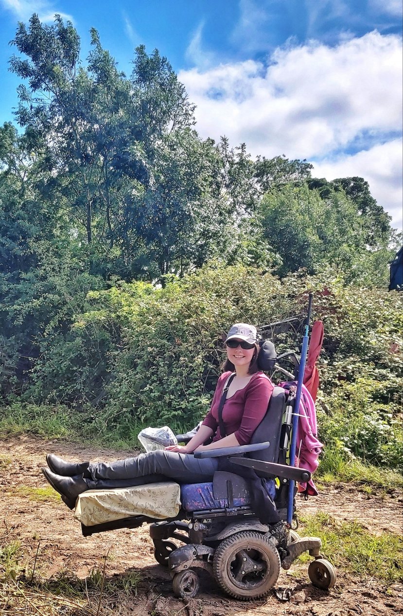 Holly, a white woman with shoulder length brown hair, sitting outside in front of hedges and trees in a Quickie Q700 Sedeo Pro Powered Wheelchair with the legs elevated. Wearing grey trousers, a plum coloured top, sunglasses and a grey baseball cap.