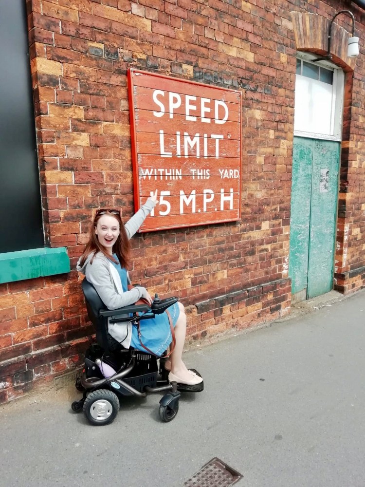 Pippa, a white woman with brown hair and wearing a blue dress and grey jacket, sitting in a powered wheelchair pointing and laughing at a sign saying "Speed limit within this yard 15 mph"