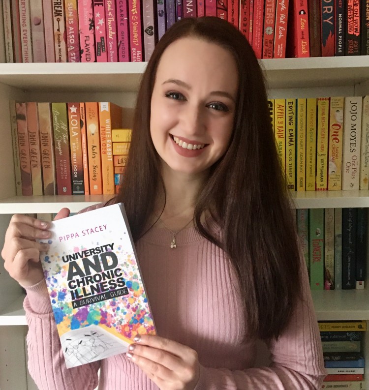 Pippa, a white woman with long brown hair, smiling in front of a bookcase with a rainbow of books while holding the book "University and chronic illness, a survival guide". 