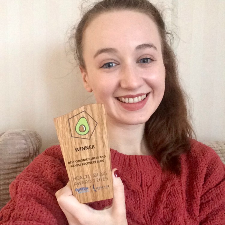 Pippa, a white woman with brown hair tied back, wearing a red jumper and smiling while holding a wooden Health Blog Awards trophy for best chronic illness and recovery blog.