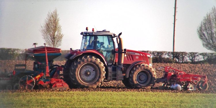 A red Massey Ferguson tractor driving across a field, with a set of discs on the front and a powerharrow and drill behind, drilling a crop.